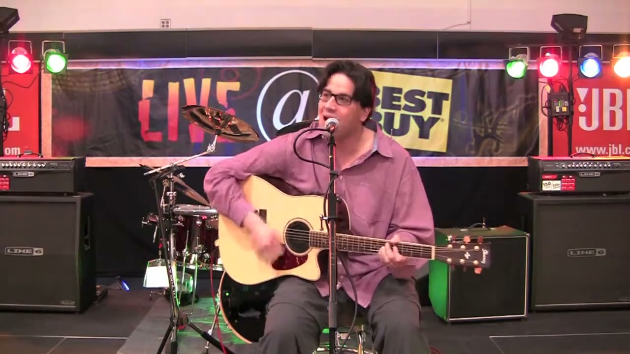 Tony seated with an acoustic guitar on a small store stage
