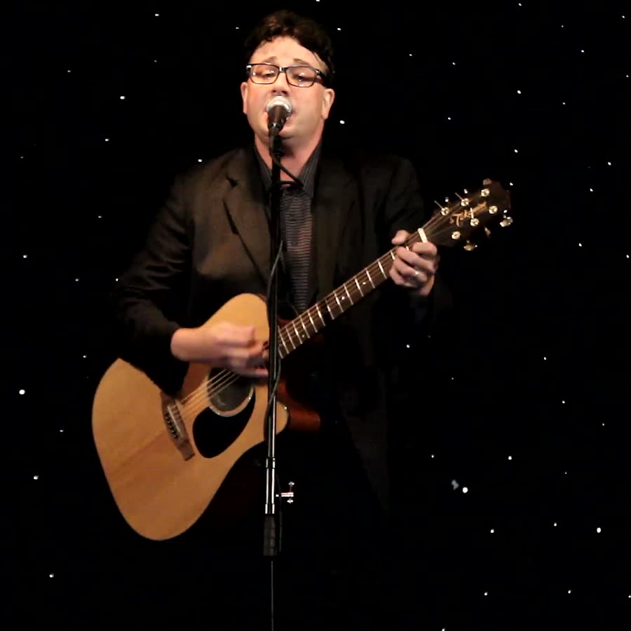 Tony Costello on stage in a dark suit and glasses, singing into a microphone and playing an acoustic guitar