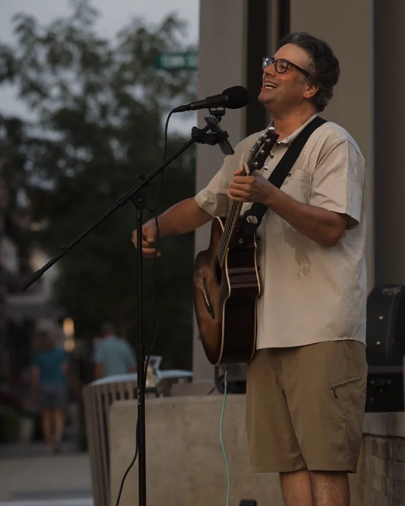 Tony Costello playing an acoustic guitar outdoors at a microphone, smiling up at the crowd
